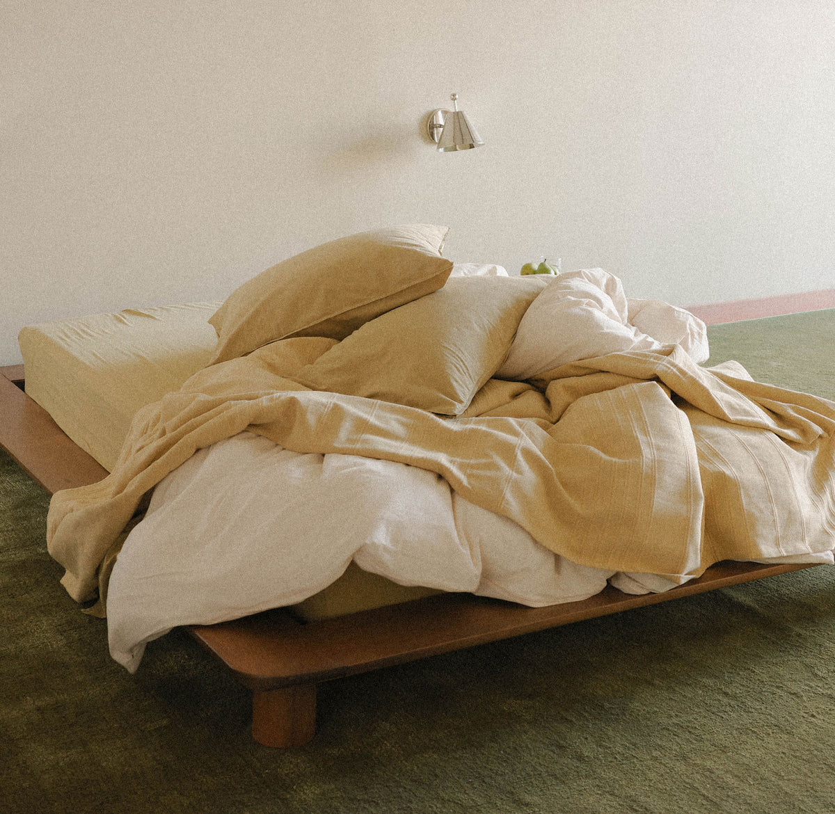 A low wooden bed dressed in soft beige and off-white bedding, with rumpled duvet and pillows, set against a plain light wall with a small wall lamp.