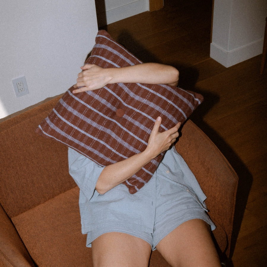 A person sitting on a brown armchair, holding a brown and purple plaid throw pillow against their face. They are wearing light blue shorts and a matching top.