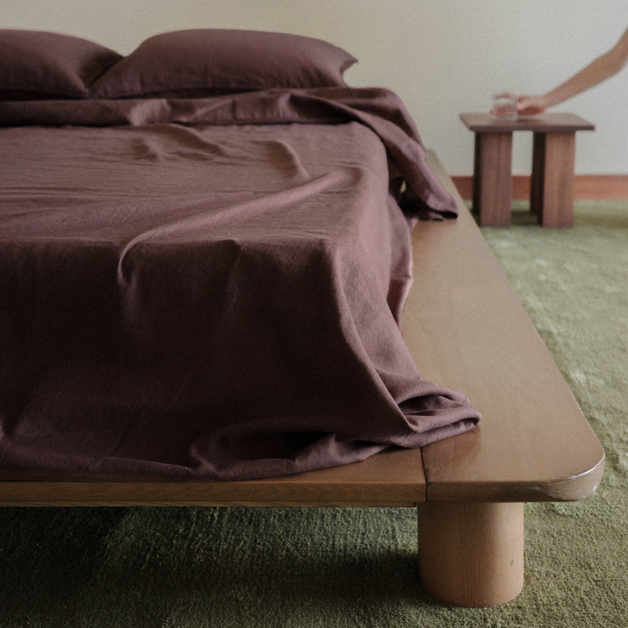 A bed dressed in dark plum-brown bedding, shown in a real bedroom setting with a wooden platform bed frame, green carpet, and a person's arm visible at the upper right adjusting a small wooden stool.