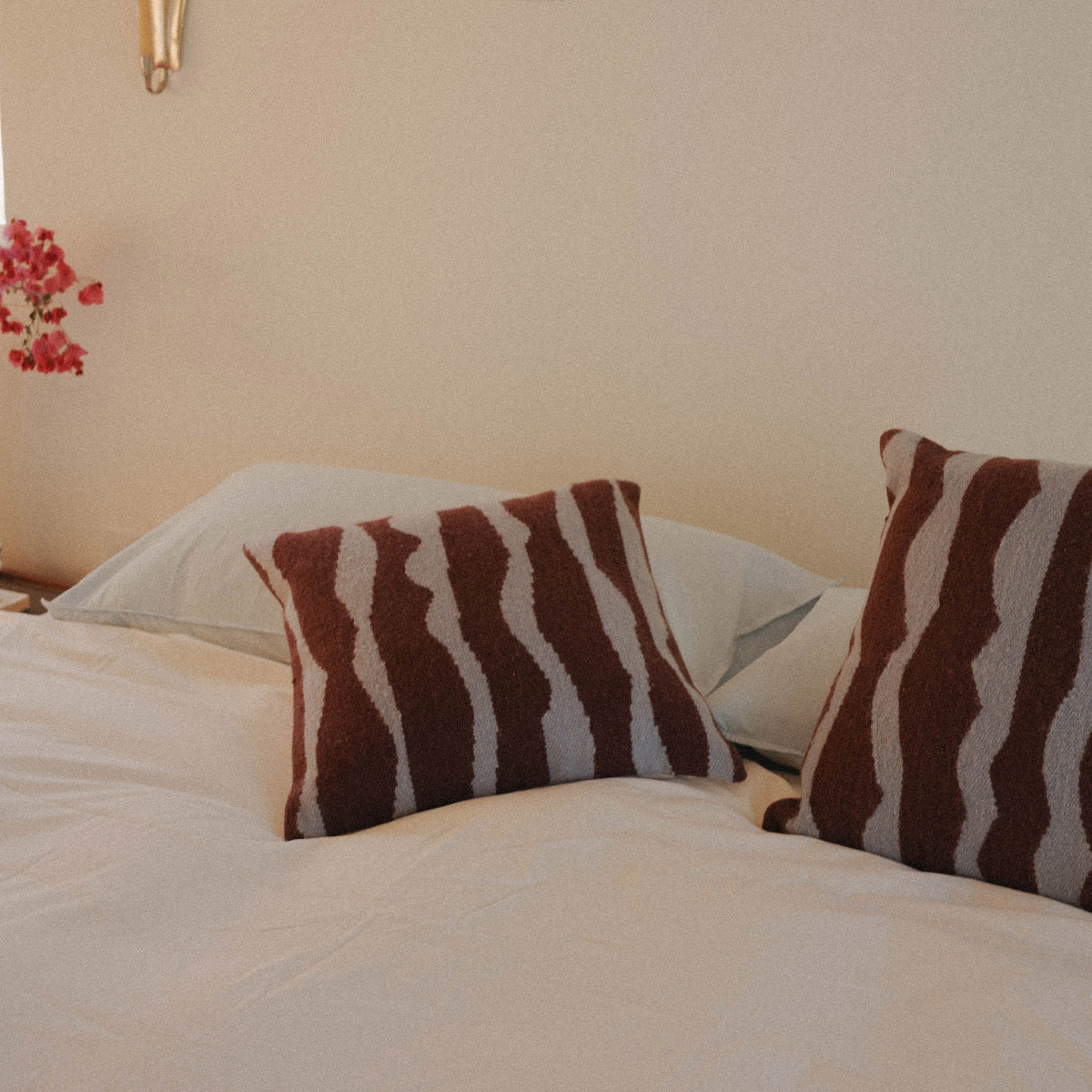 A bed dressed in light blue sheets, cream duvet with two brown and blue organic striped pillows, positioned in a warmly lit bedroom.