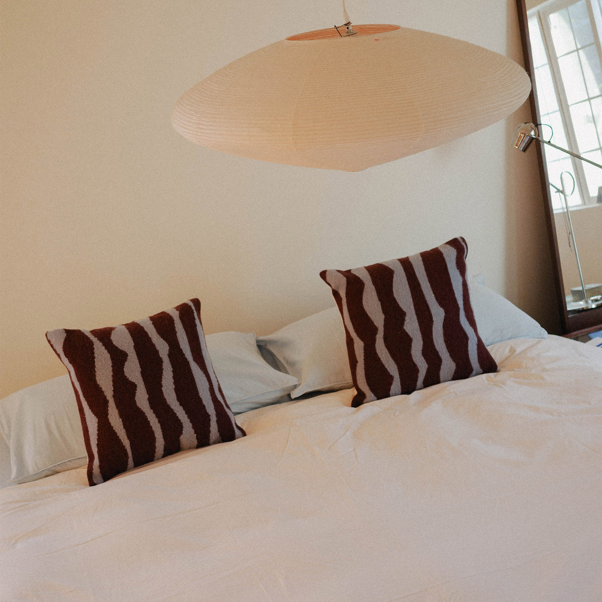A bed dressed in light blue sheets, cream duvet with two brown and blue organic striped pillows, positioned in a warmly lit bedroom.