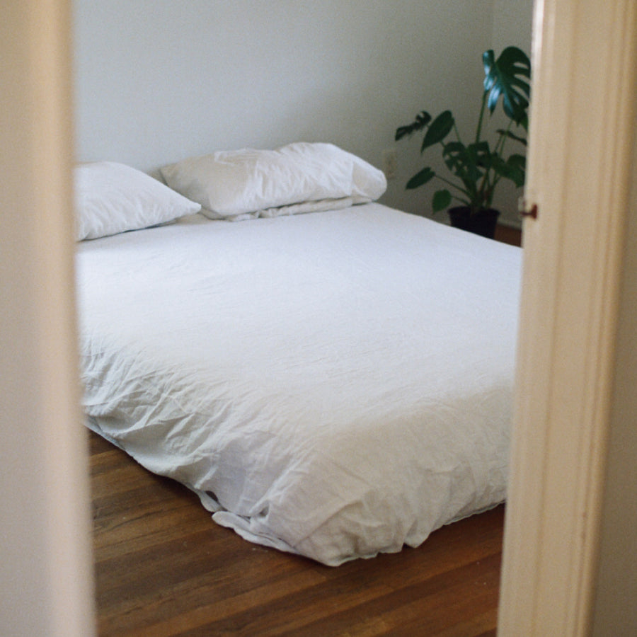 A neatly made bed with white bedding and two pillows, photographed from an open doorway in a bedroom with wood flooring and a potted plant in the corner.
