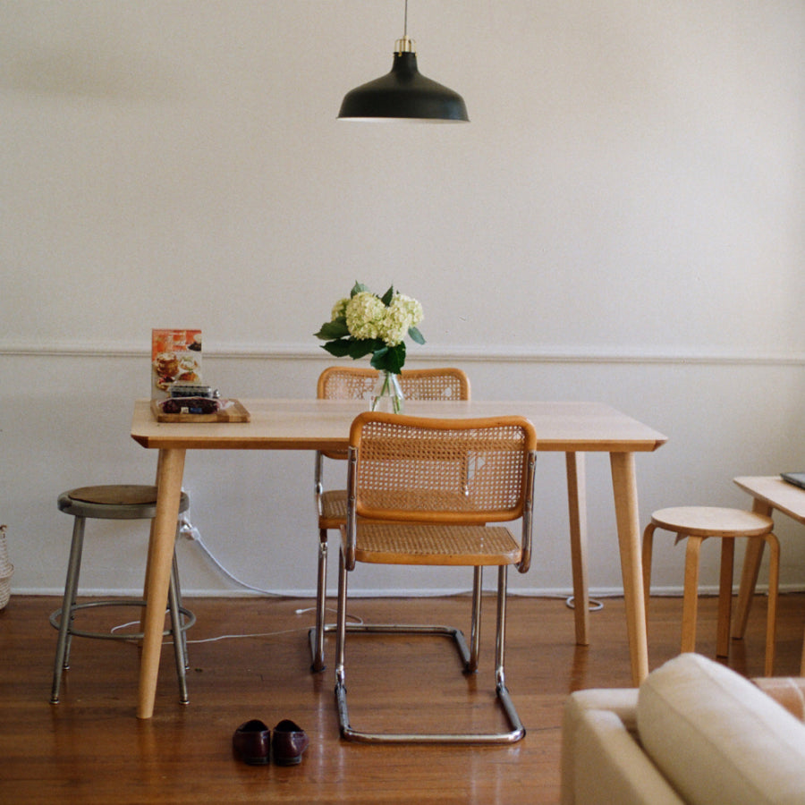 A light wood dining table with four woven cane chairs and a black pendant light hanging above, set in a minimalist room with a white wall and wooden floor.