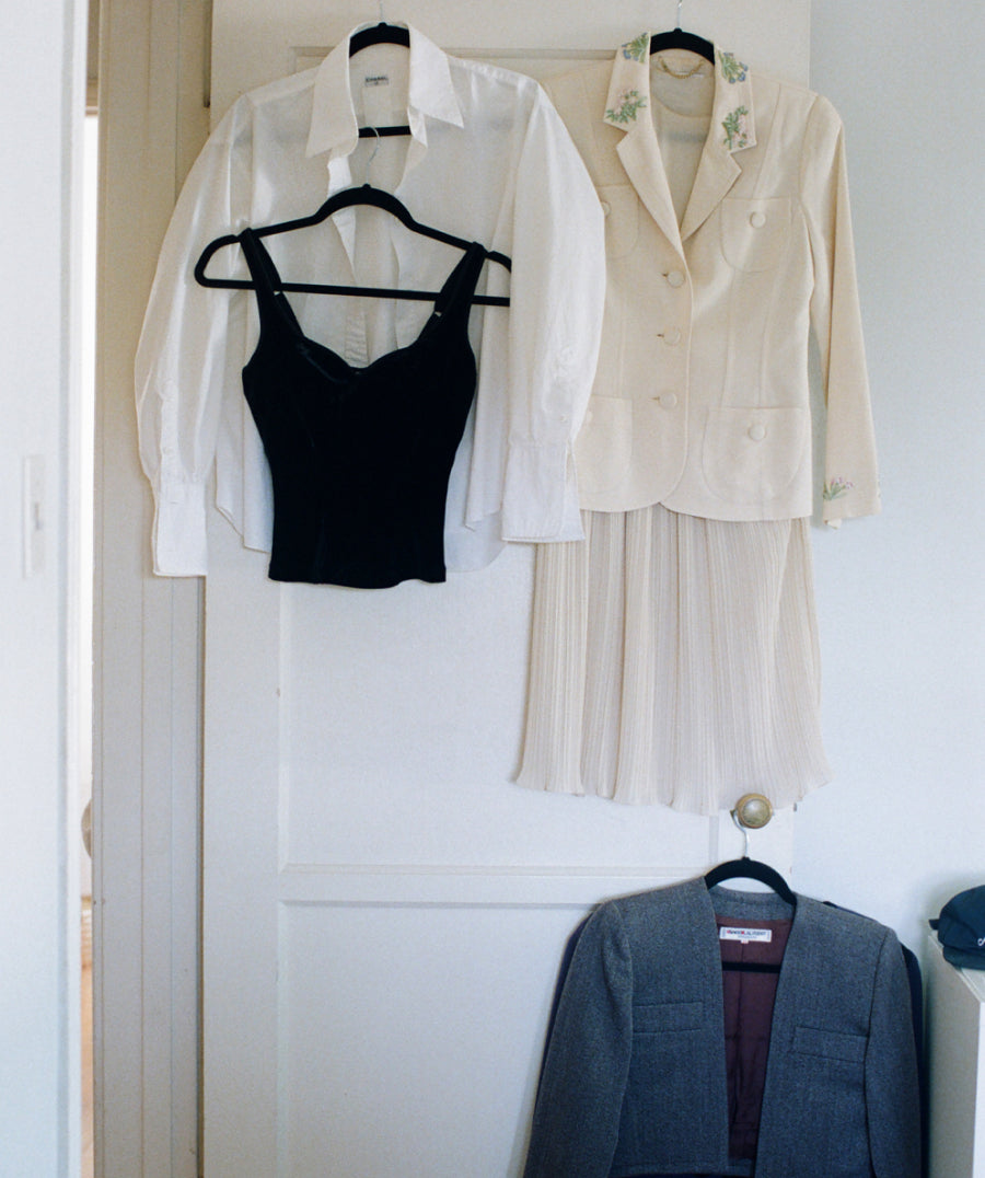 Two white button-up shirts, a black camisole, and a pleated cream skirt are displayed on hangers against a white paneled door, with a gray blazer hanging below.