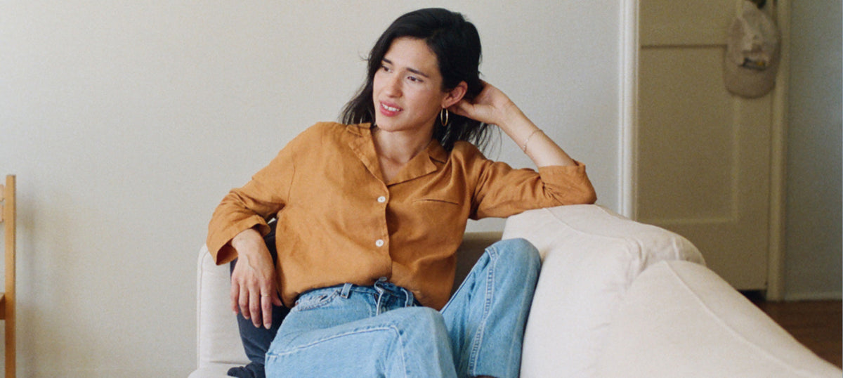A woman sitting on a white armchair in a minimalist room, wearing a camel button-up shirt and light blue jeans.