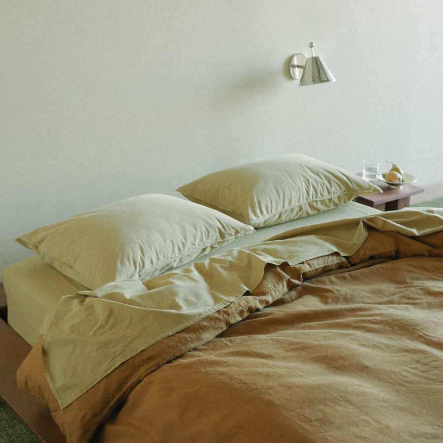 A neatly made bed with matching beige pillows and sheets, topped with a green-brown duvet cover in a softly lit bedroom. A small bedside table with a glass and fruit sits against a plain wall with a wall lamp.