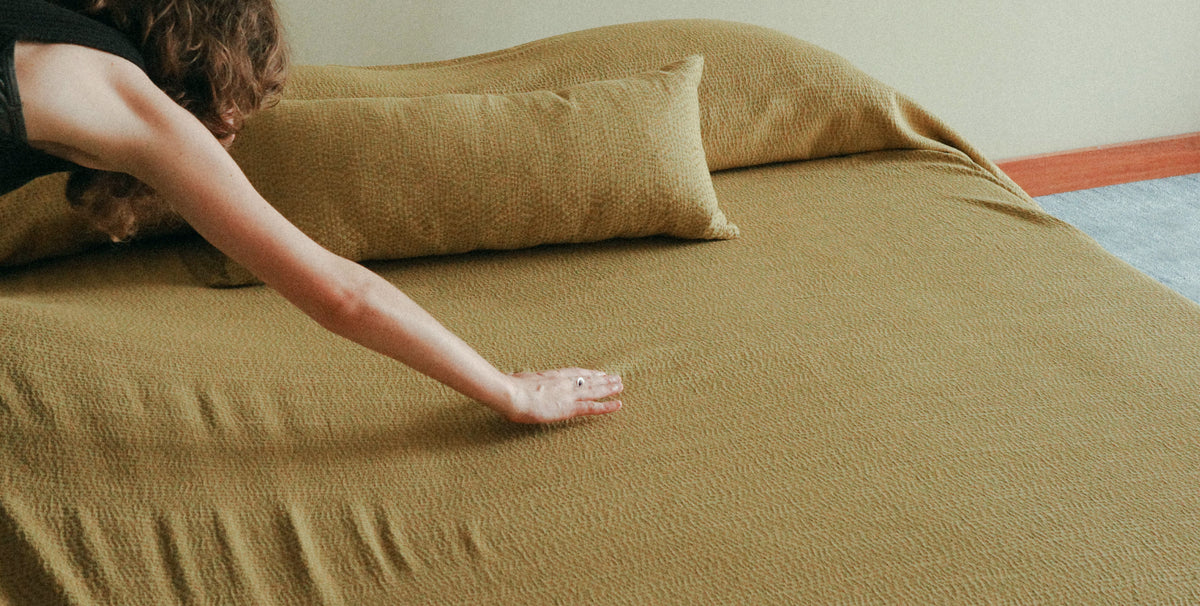 A person's arm and hand stretching a textured olive green bedspread over a bed with matching pillows in a bedroom setting.