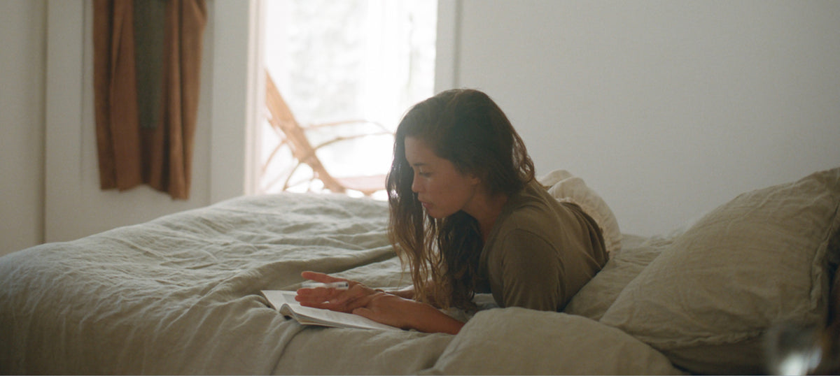 Woman laying on a bed writing in a journal on top of sage green linen bedding in a bright bedroom