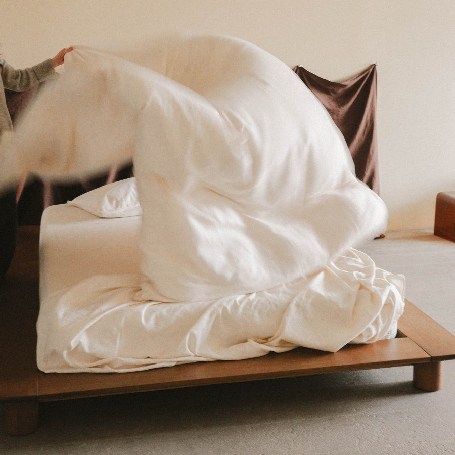 A person's hand and arm are visible as they lift and spread a large, soft white duvet cover or comforter over a bed on a wooden platform bed frame in a minimalist bedroom setting.