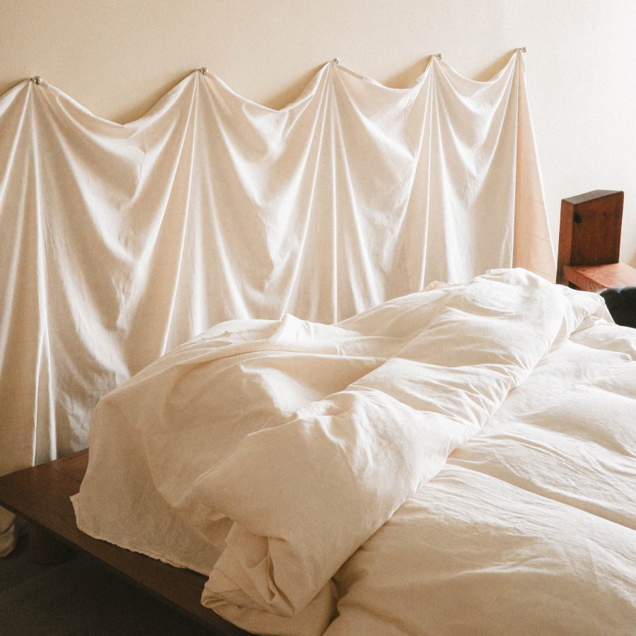 A bed dressed in soft off-white bedding, with a matching draped fabric backdrop hanging behind it in a softly lit room.