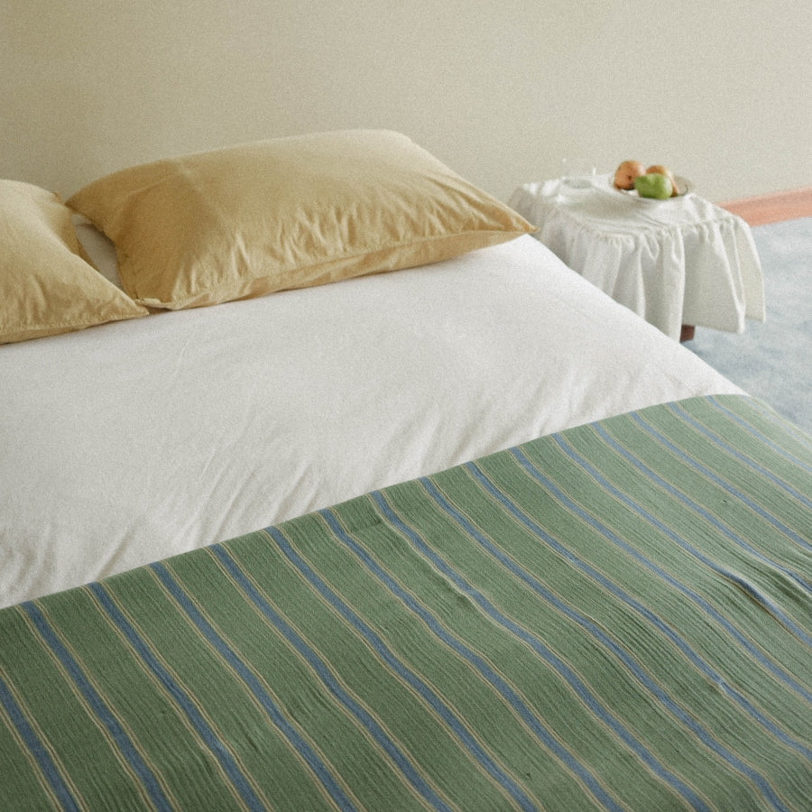 A bed dressed in white bedding with two light yellow pillows and a green striped blanket draped across the foot, set in a softly lit room with a small table holding fruit in the background.