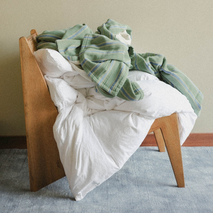 A wooden armchair with a white duvet and a green striped blanket draped over it, set against a plain beige wall and light blue-gray carpet.