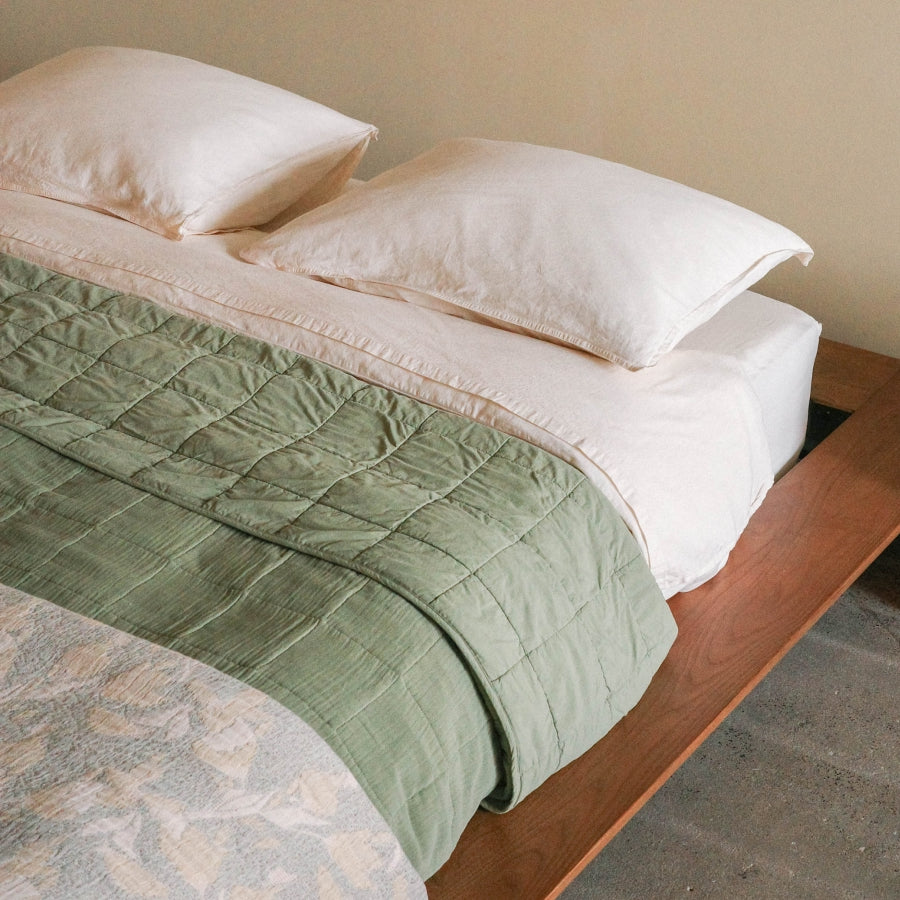 A neatly made bed with two cream pillows, and a juniper green quilted blanket draped over the foot of a wooden bed frame in a neutral room.