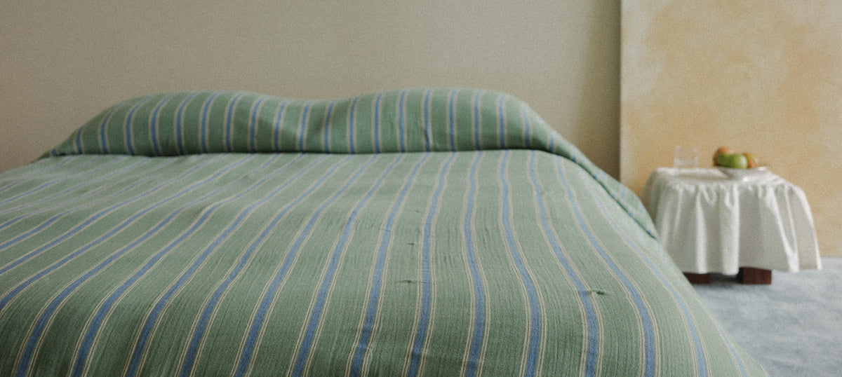 A bed made with a green striped duvet cover and matching pillowcases, set in a simple bedroom with a small side table holding a glass and fruit.