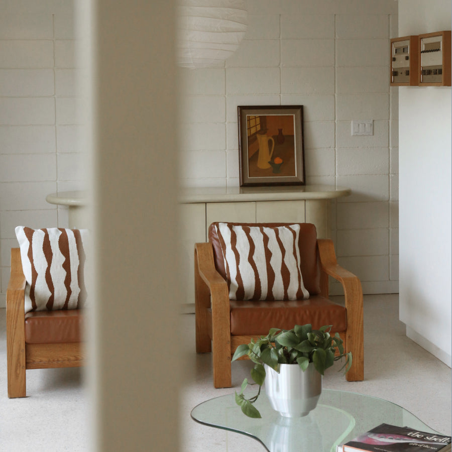 A mid-century modern living room scene featuring two wooden armchairs with brown leather seats and white pillows with brown wavy stripes. A glass coffee table with a potted plant is in the foreground, and a tiled wall with a framed picture is in the background.