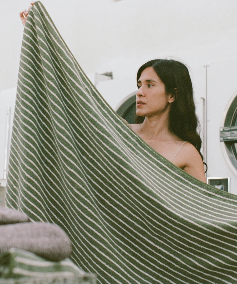 A woman holds up a large striped blanket in front of her. The fabric has a dark olive green base with thin white stripes.