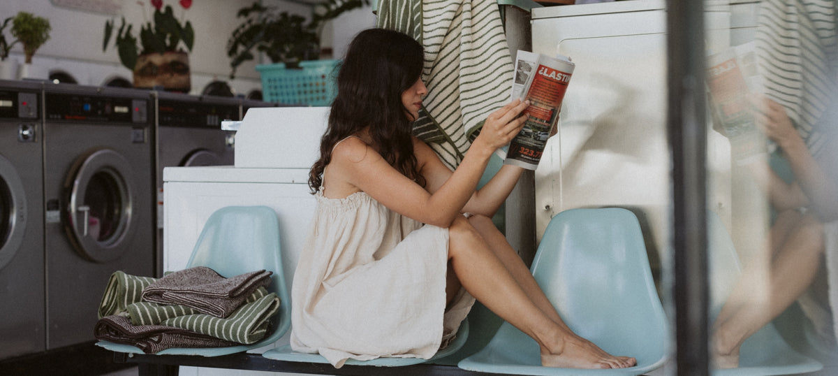 A woman in a light beige dress sits on a light blue chair in a laundry room, reading a magazine. 
