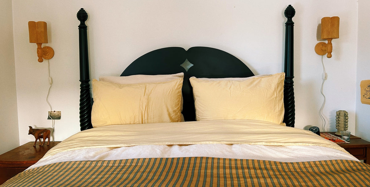 A bed with cream and yellow bedding, and a striped yellow-and-brown blanket, set against a white wall with wooden wall lamps and bedside tables.