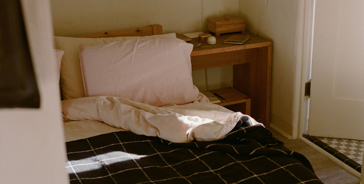 An unmade bed with pink pillowcases and a black check blanket peeks out from behind a white wall in a warmly lit room