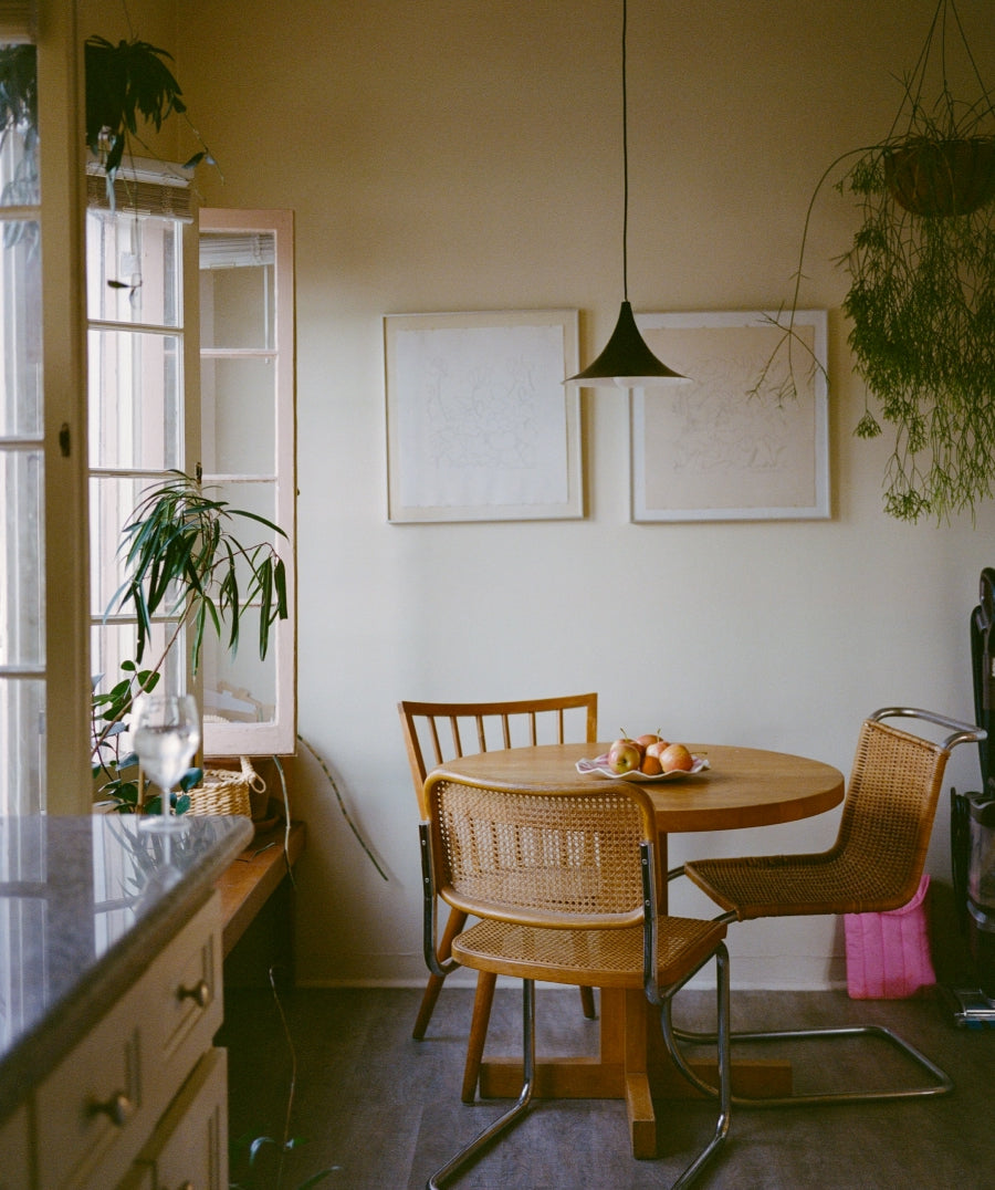 A cozy interior corner with a round wooden dining table and two woven wicker chairs, set against a warm beige wall with two blank framed prints. A black pendant light hangs above, and a window on the left with open panes lets in natural light, accompanied by potted plants.