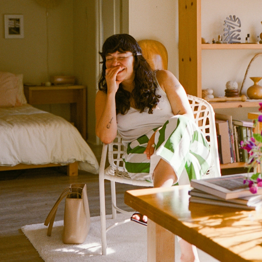 A woman sits in a white chair in a sunlit bedroom, wearing a white sleeveless top and green-and-white striped shorts. A beige handbag rests on the floor beside her, with a bed, shelves, and a wooden table visible in the room.