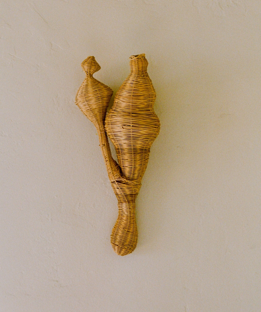 A natural woven rattan wall hanging sculpture shaped like two abstract female figures, mounted on a textured beige plaster wall.