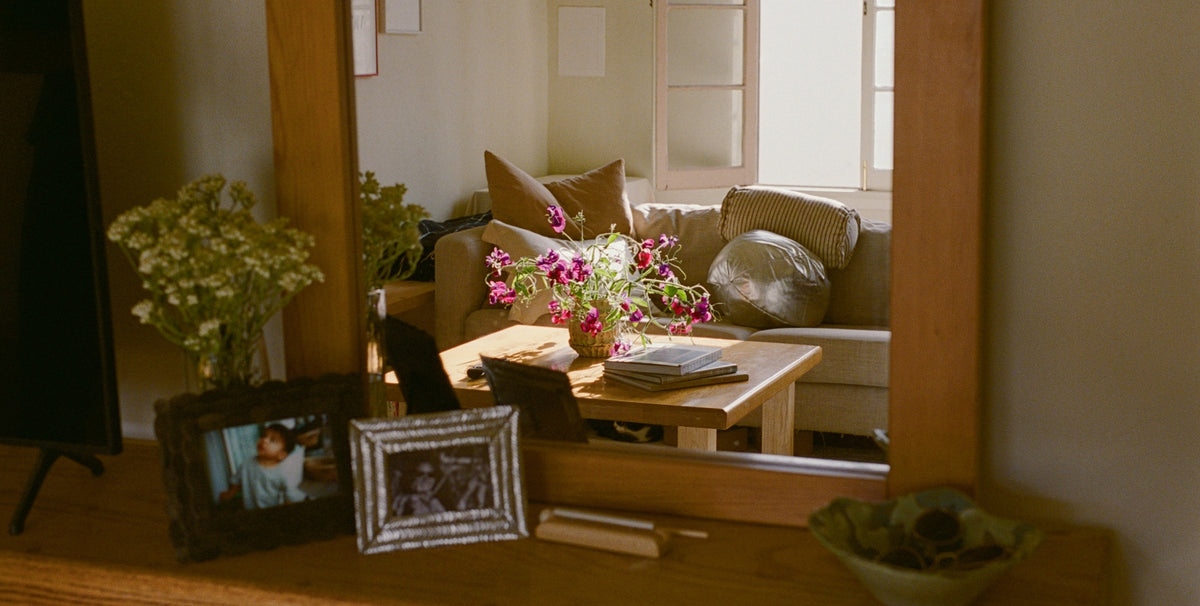 A warm, cozy interior scene with a wooden-framed mirror reflecting a living room setup with a sofa, pillows, flowers, and a coffee table. In the foreground, a wooden surface holds framed photos, a vase of flowers, and a bowl.