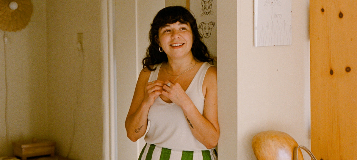 A smiling woman stands in a doorway wearing a white tank top and green-and-white striped bottoms, with a warm indoor setting behind her.
