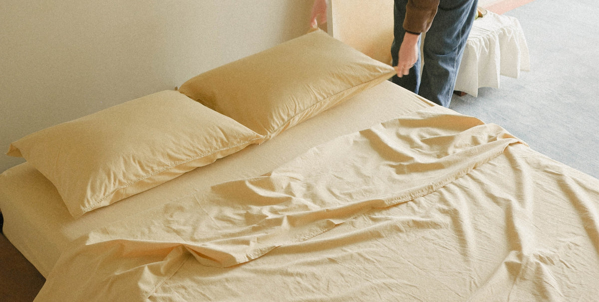 A bed dressed in light yellow bedding, including two pillows and a rumpled sheet, with a person's hands and legs visible adjusting the top corner of the duvet in a softly lit room.