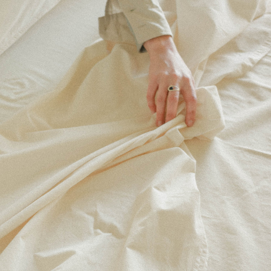 A close-up of a person's hand adjusting cream-colored bedding on a bed, with a matching long-sleeve shirt partially visible.