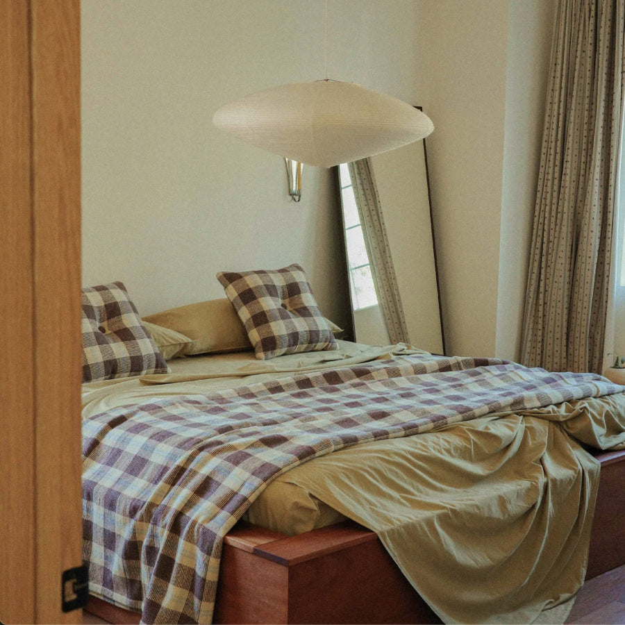A cozy bedroom scene featuring a bed dressed in beige and brown plaid bedding, and matching pillows with a wooden headboard and a tall floor mirror in the background.