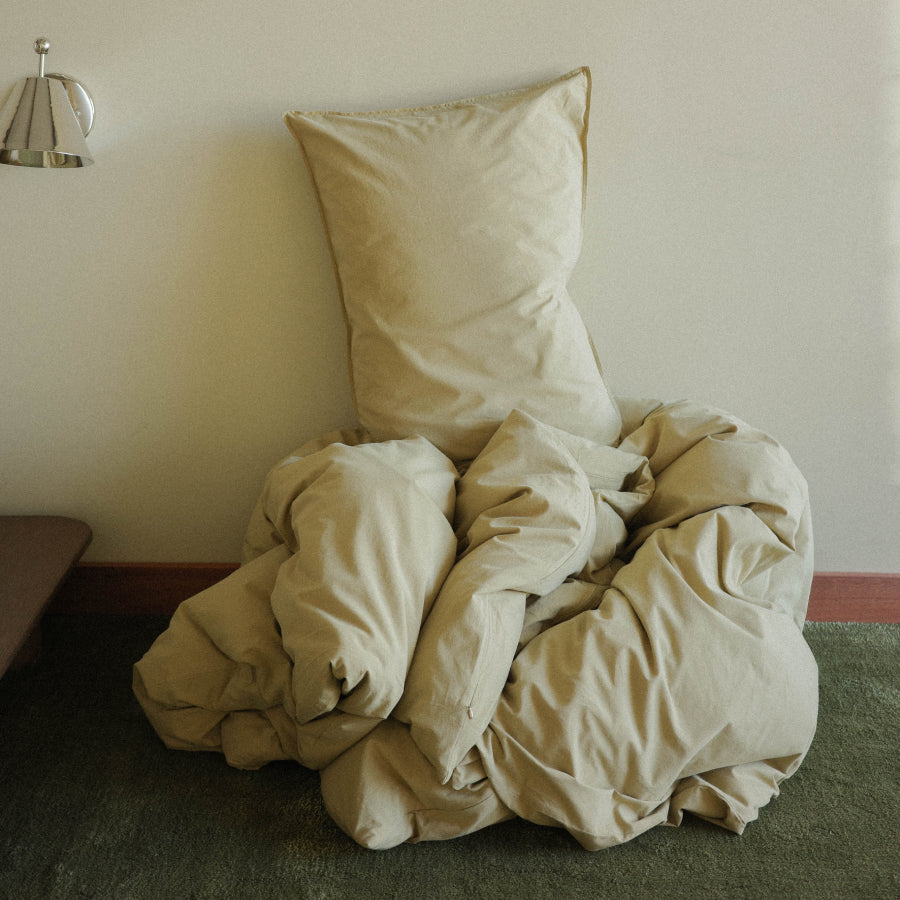 A light green pillow and matching rumpled bedding or duvet arranged on a dark green carpet against a plain off-white wall.