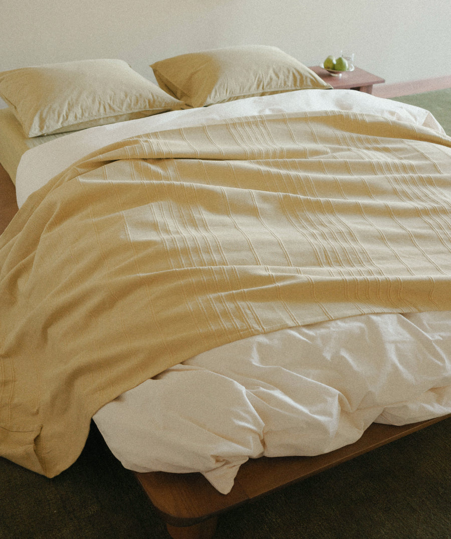 A cozy bed styled with a mustard yellow textured throw blanket layered over white sheets and duvet, with two matching beige pillows, in a softly lit bedroom setting.