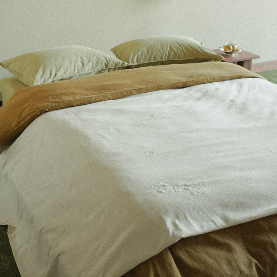 A neatly made bed with two contrasting green-colored bedding and a warm beige throw blanket, styled in a minimalist bedroom setting with matching pillows and a small wooden bedside table.