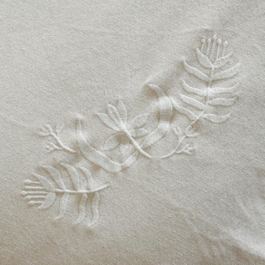 A close-up of a light beige fabric with a subtle, tonal embroidered botanical pattern of leaves and vines.