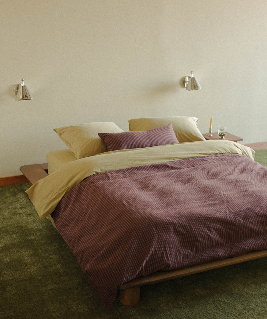 A bed dressed in light green and burgundy striped bedding, set in a minimalist bedroom with a wooden platform bed frame, green carpet, and wall-mounted sconces.