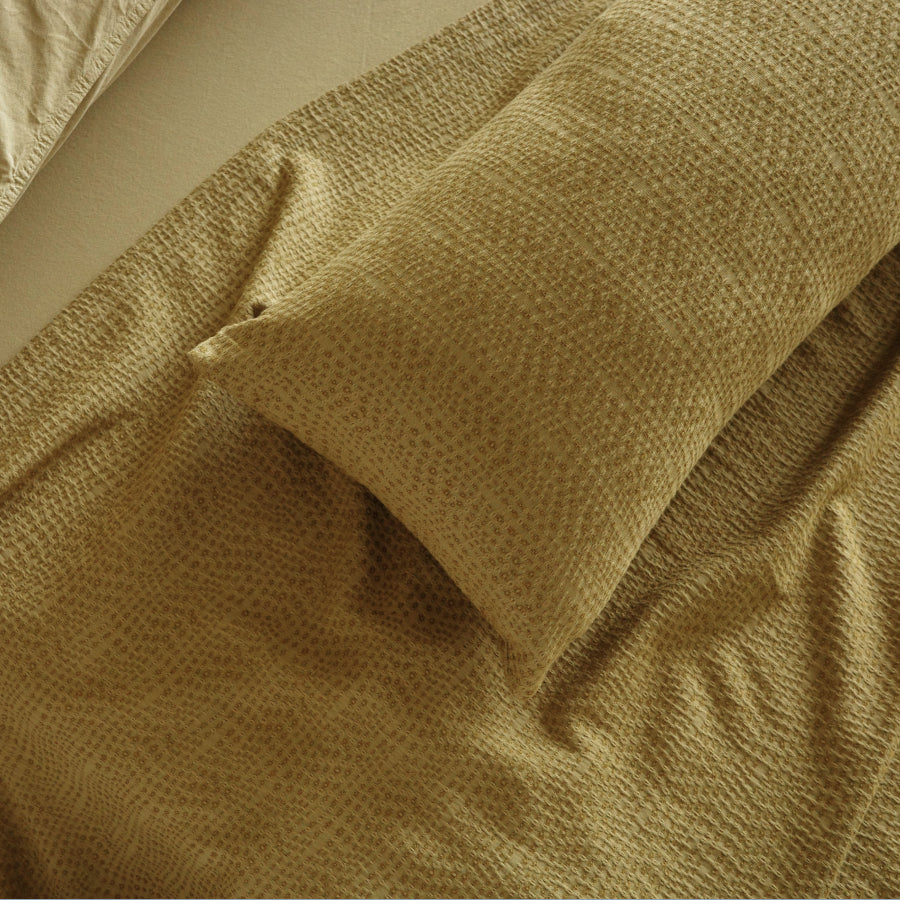 A close-up of a textured waffle-weave bedding set in a warm green color, featuring a pillowcase and matching sheet.