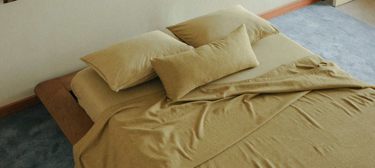 A bed dressed in matching olive-khaki bedding, including a sheet, pillowcases, and a folded top sheet, styled in a bedroom setting with a blue carpet and light-colored wall.