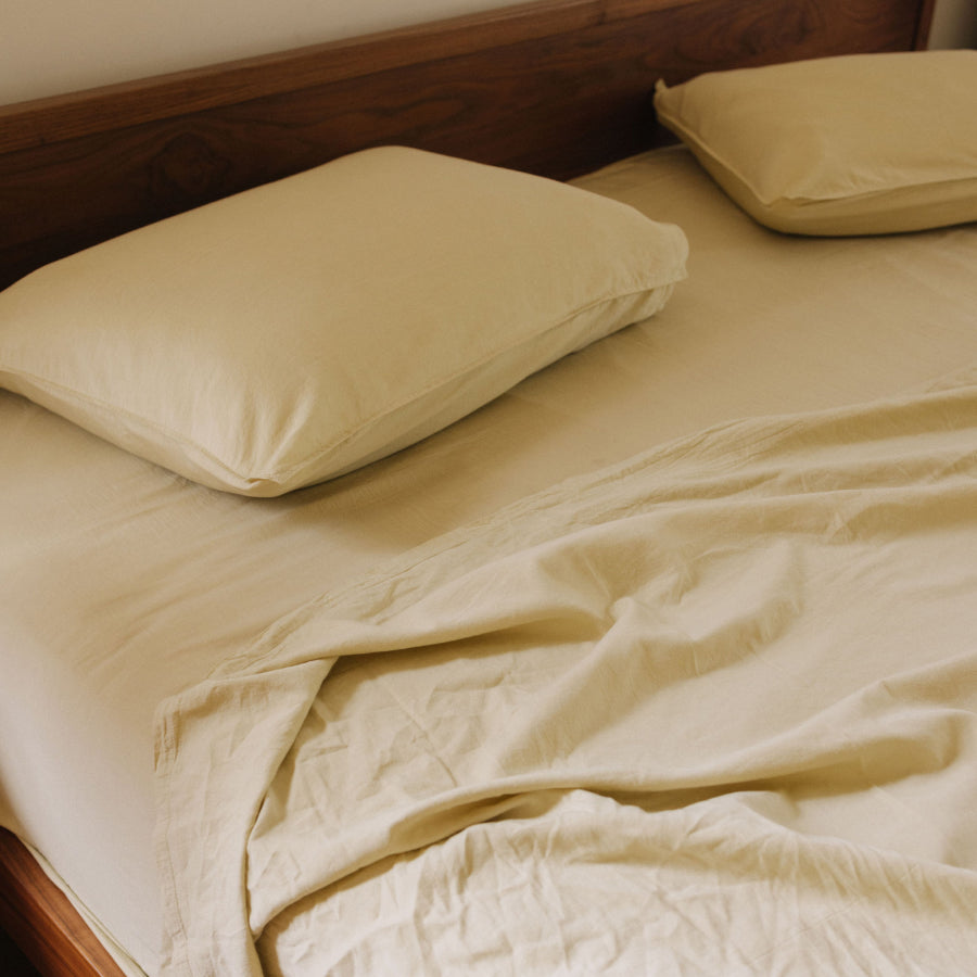 Light yellow cotton bed sheets and matching pillowcases on a bed with a dark wood headboard.