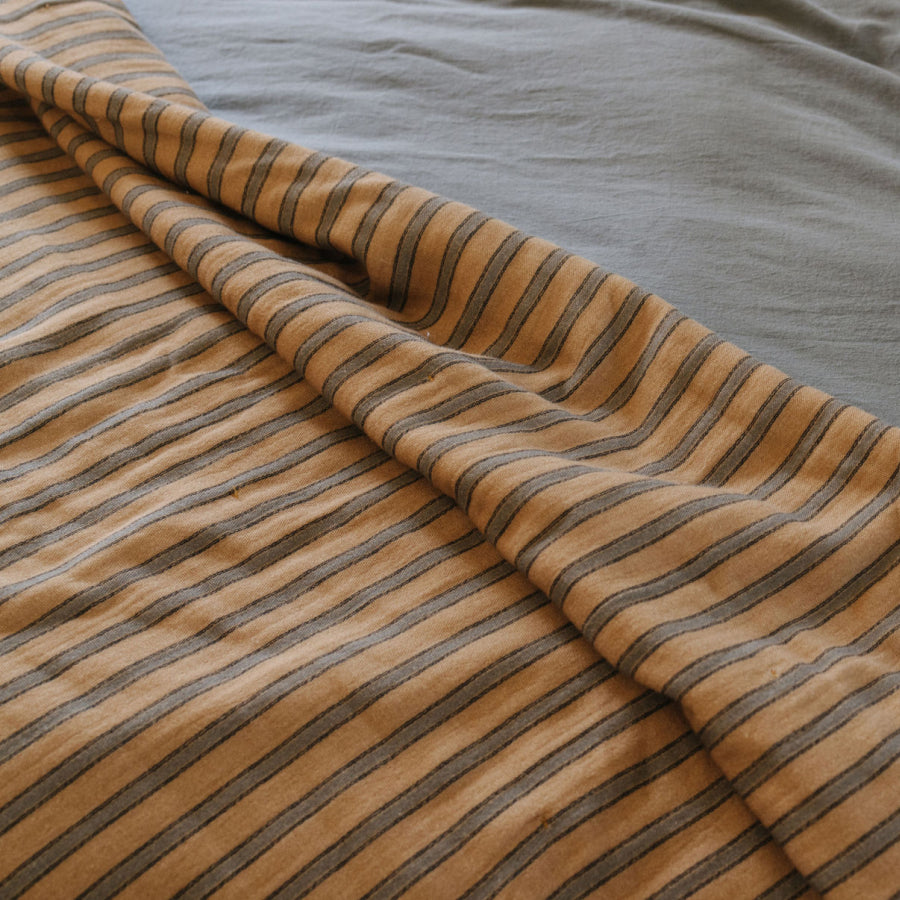 A close-up of a striped blanket draped over a blue green sheet. The fabric features alternating stripes of warm tan/beige and dark brown.