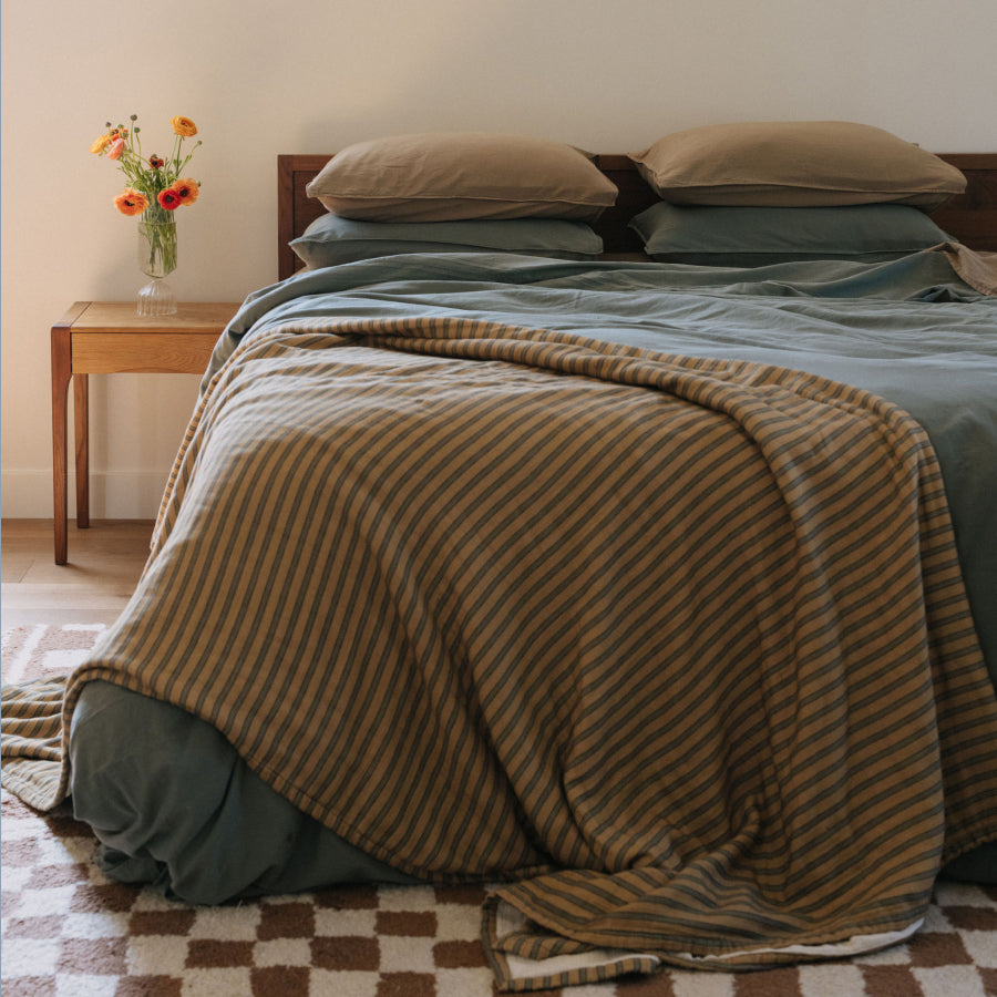 A cozy bedroom scene with a bed made up in muted sage green bedding and a brown-and-tan striped throw blanket draped over the foot of the bed. 