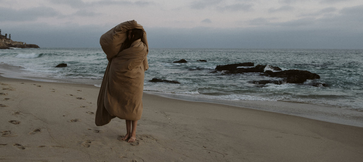 A person wrapped in a large tan blanket stands on a sandy beach under an overcast sky, with rocks and waves in the background.