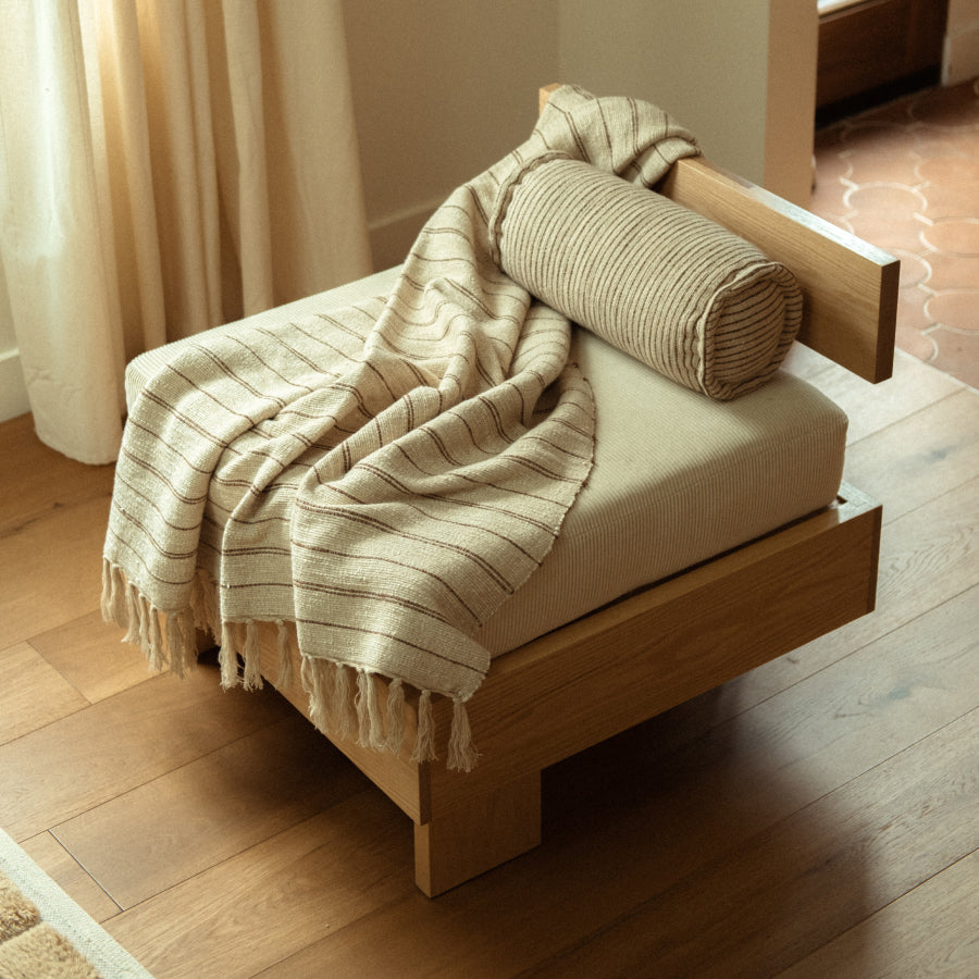 A low wooden chair with a cream cushion, draped with a beige striped fringed throw blanket and topped with a matching bolster pillow, set in a softly lit room with wood flooring and curtains.