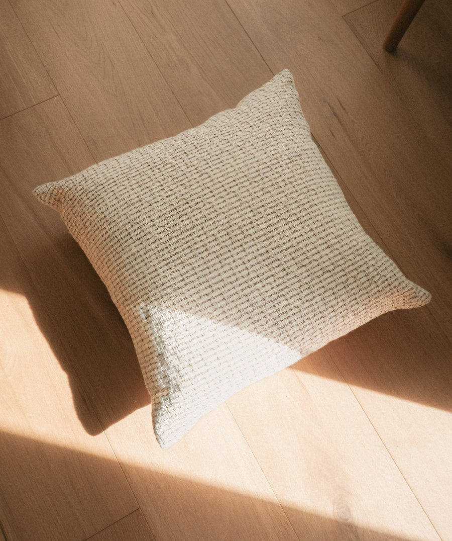 A square throw pillow with a textured, woven waffle-like pattern in a light cream color, resting on a light wood floor with natural sunlight casting shadows.
