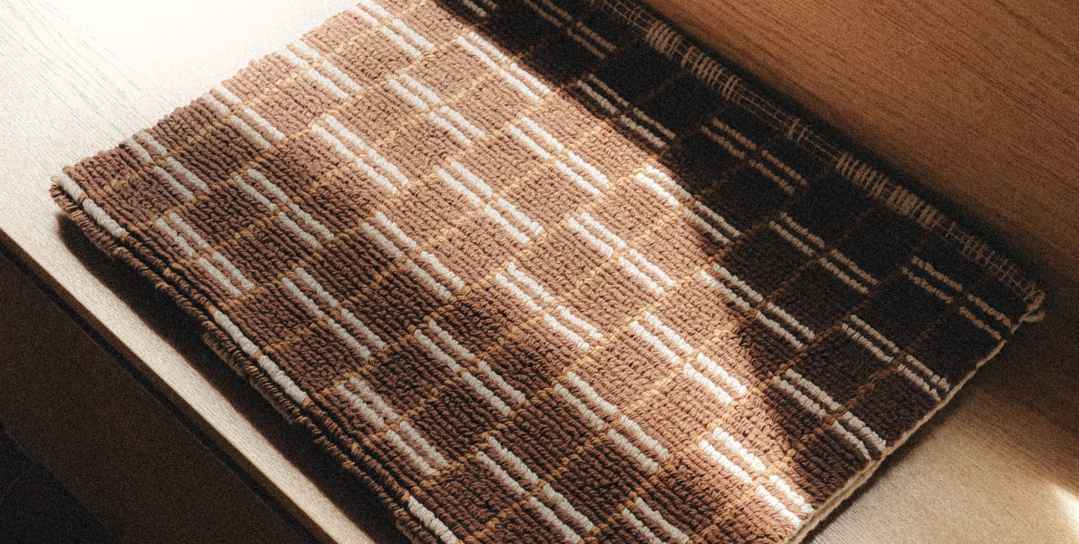A close-up of a woven brown and beige bath mat laid on a light surface with warm sunlight and shadows.