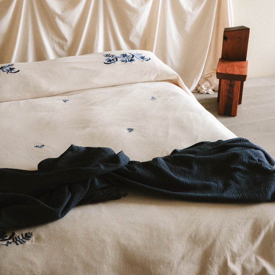 A bed dressed in light beige bedding with small blue floral embroidery, styled with a dark navy textured throw blanket and a small wooden stool in the background.