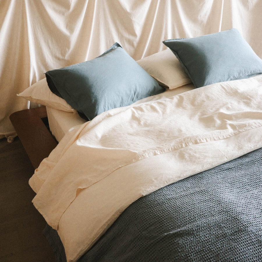 A neatly made bed with off-white bedding, two muted blue pillows, and a textured blue blanket, photographed in a softly lit room with draped fabric in the background.