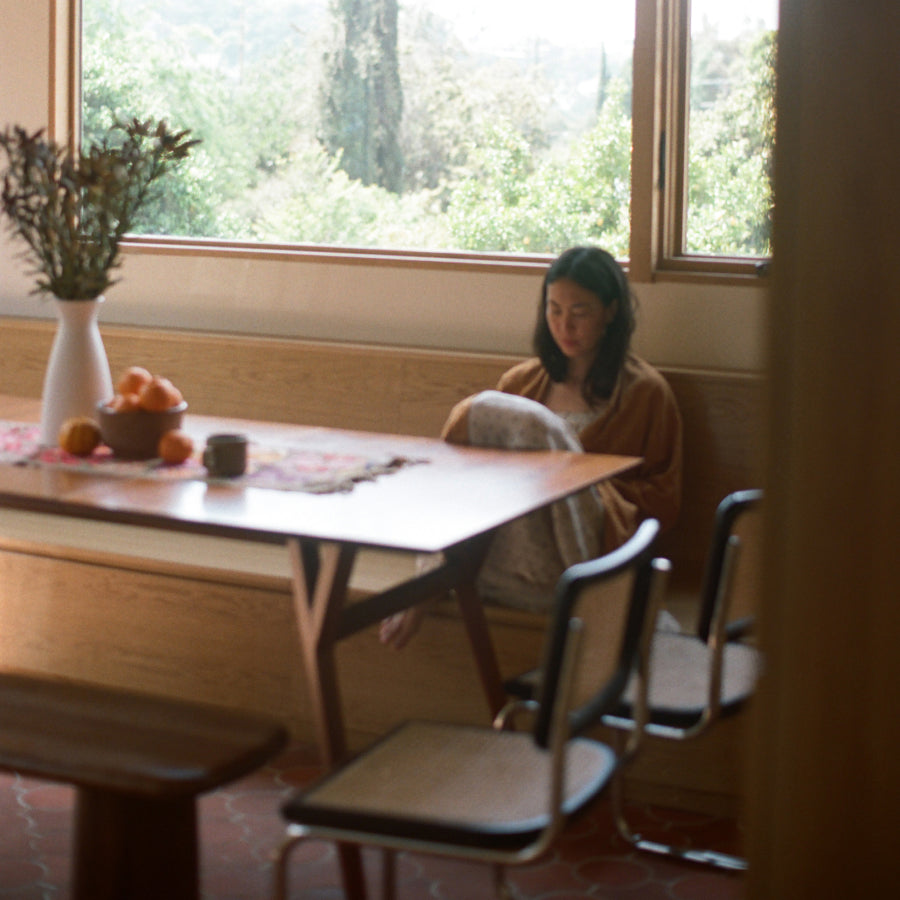 A woman sits at a wooden dining table in a sunlit room, wearing a robe and sleepdress. The scene includes chairs, a vase with dried stems, oranges on the table, and large windows showing greenery outside.
