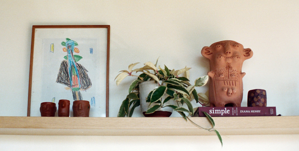 A terracotta-colored ceramic monkey figurine with carved details sits on a wooden shelf next to a potted variegated plant and a framed abstract drawing on a white wall.