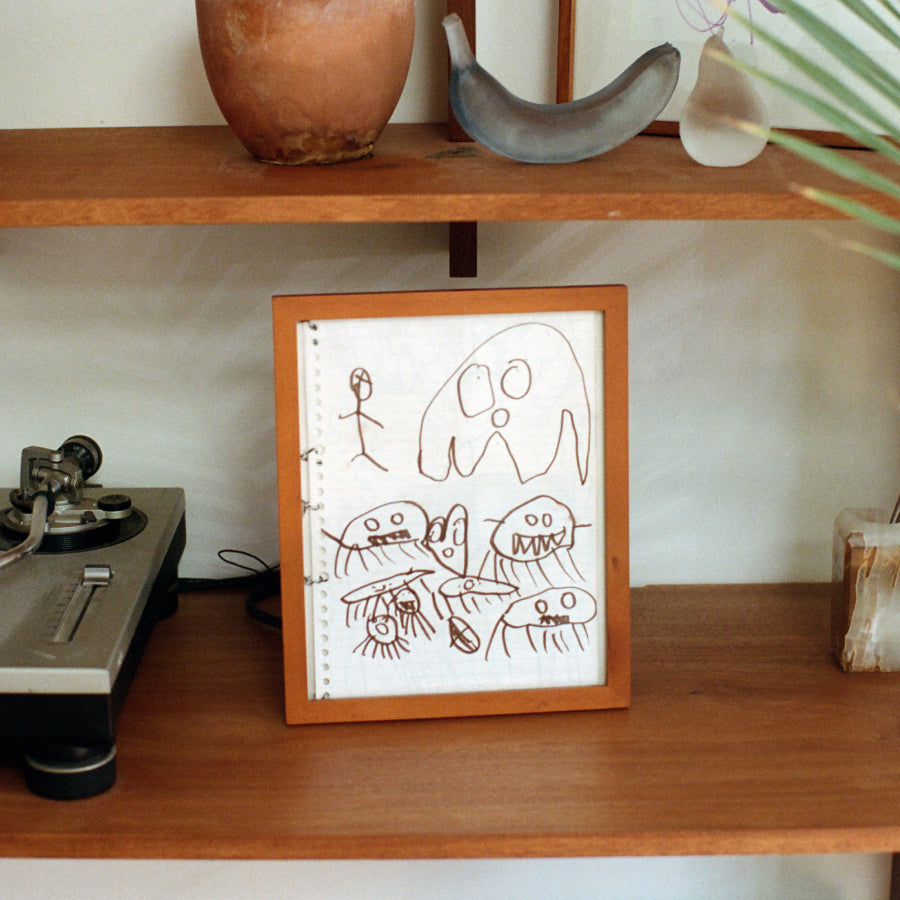 A framed childlike drawing on white paper with brown scribbled figures, displayed on a wooden shelf next to a turntable and other home decor.